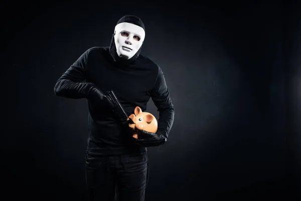 Burglar in mask holding gun and piggy bank — Stock Photo