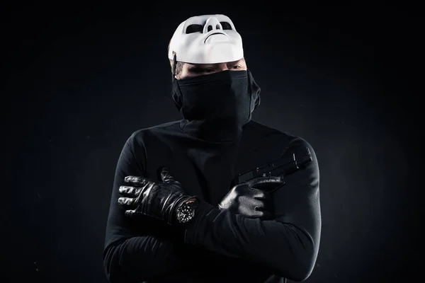 Thief in black balaclava and white mask holding gun in folded arms on black — Stock Photo