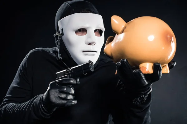 Robber in balaclava and mask aiming at piggy bank — Stock Photo