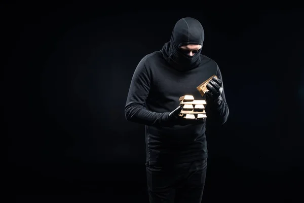 Thief in balaclava stacking gold ingots in his hands — Stock Photo