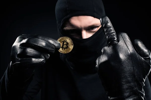 Man in black balaclava holding golden bitcoin and showing middle finger — Stock Photo