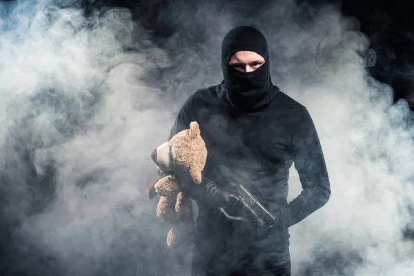 Criminal em balaclava segurando arma e ursinho de pelúcia em nuvens de fumaça — Fotografia de Stock