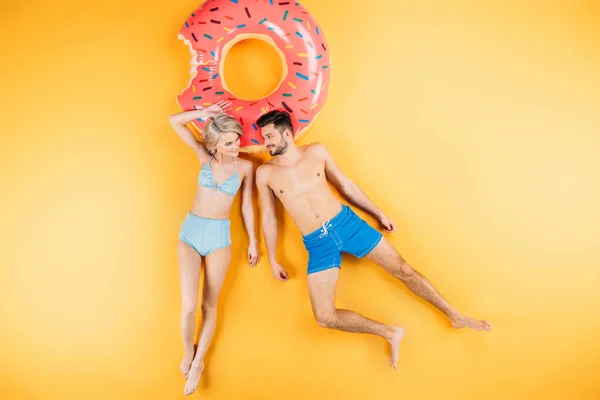 Feliz joven pareja en traje de baño sonriendo el uno al otro mientras está acostado en el anillo de natación en amarillo - foto de stock