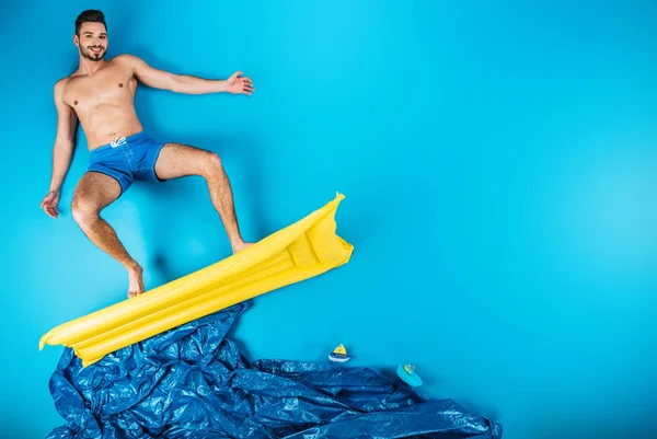 Schöner junger Mann in kurzen Hosen lächelt in die Kamera, während er auf einer aufblasbaren Matratze auf blauem Grund steht — Stockfoto