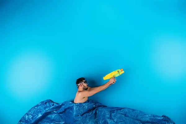 Young man in swimming mask shooting with water gun from imagine waves on blue — Stock Photo