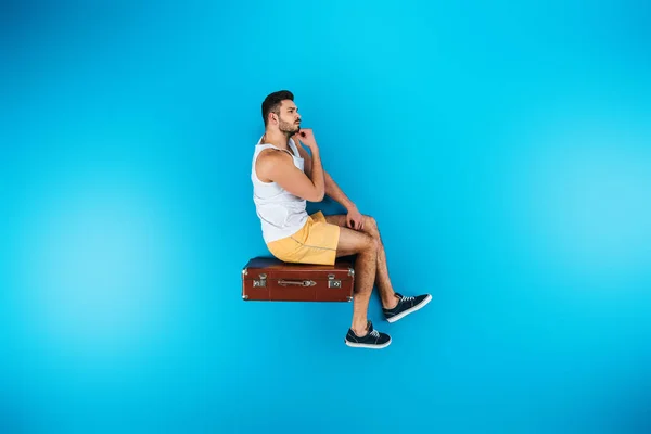 Schöner junger Mann sitzt auf Koffer auf blauem Sommerferienkonzept — Stockfoto