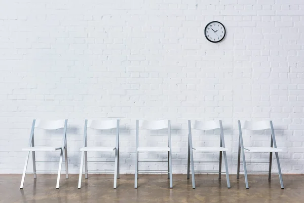 Ряд пустых стульев по белой кирпичной стене с часами — стоковое фото