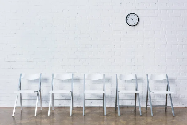 Row of empty chairs by white brick wall with clock — Stock Photo