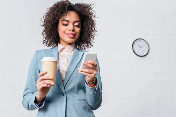 Empresaria afroamericana sosteniendo taza de café y usando smartphone - foto de stock