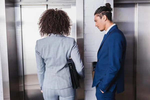 Mujer de negocios afroamericana con maletín y hombre de traje de pie por ascensor - foto de stock
