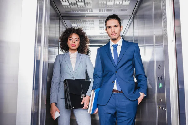 Mujer de negocios afroamericana con maletín y hombre con carpetas montando un ascensor - foto de stock
