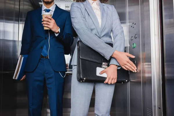 Nahaufnahme einer Geschäftsfrau, die im Fahrstuhl von einem Mann mit Kopfhörer und Kaffeetasse auf Knopf gedrückt wird — Stockfoto