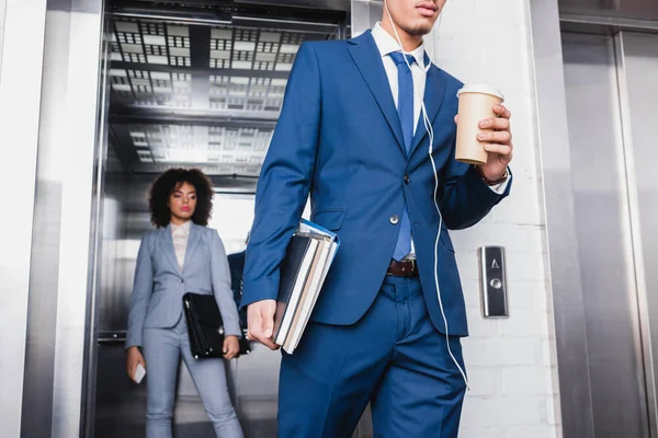 Businessman in earphones with coffee cup walking from elevator with businesswoman — Stock Photo