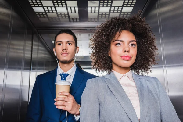 African american man in earphones with coffee cup standing in elevator with young girl — Stock Photo