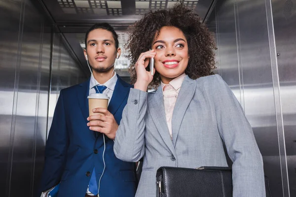 Hombre de negocios en auriculares con taza de café de pie en ascensor con mujer hablando por teléfono - foto de stock