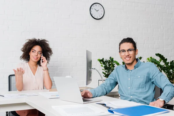 Sonriente hombre de negocios que trabaja en el ordenador portátil mientras que la mujer de negocios hablando por teléfono - foto de stock