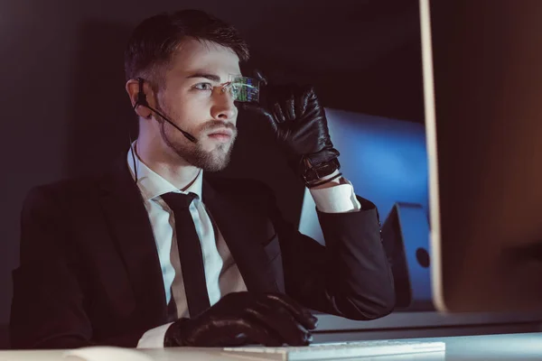 Retrato de hacker com fone de ouvido olhando para a tela do computador à mesa no escuro — Fotografia de Stock