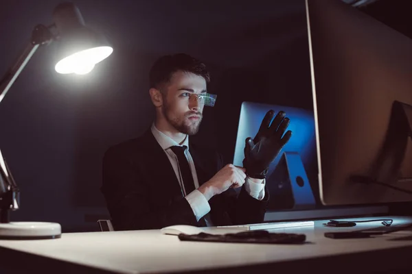 Retrato de hacker usando guantes mientras mira la pantalla de la computadora en la mesa en la oscuridad - foto de stock