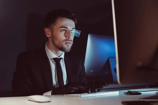 Portrait of spy agent in gloves typing and looking at computer screen at table in dark — Stock Photo