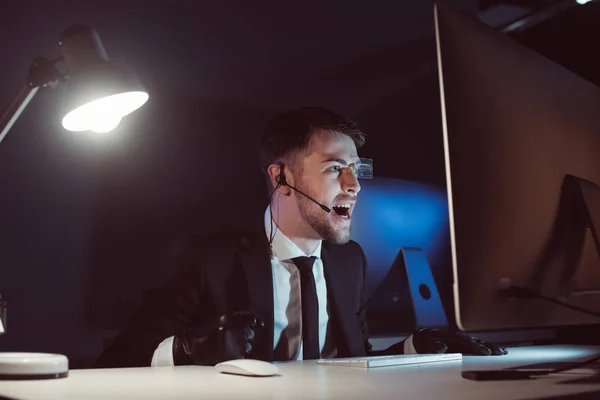 Emotional agent with headset screaming while looking at computer screen in dark — Stock Photo