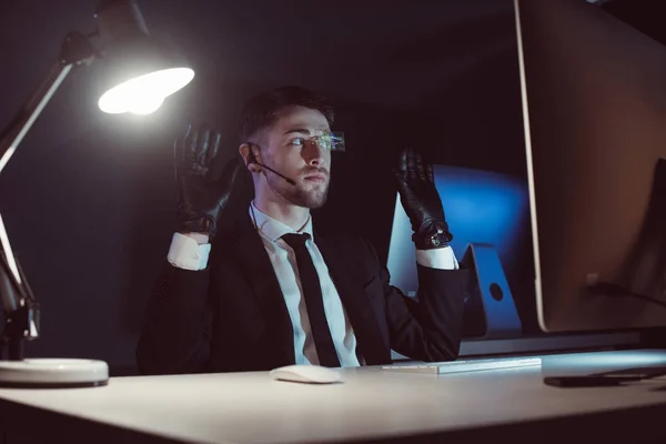 Espía en guantes con las manos arriba sentado en la mesa en la oscuridad - foto de stock