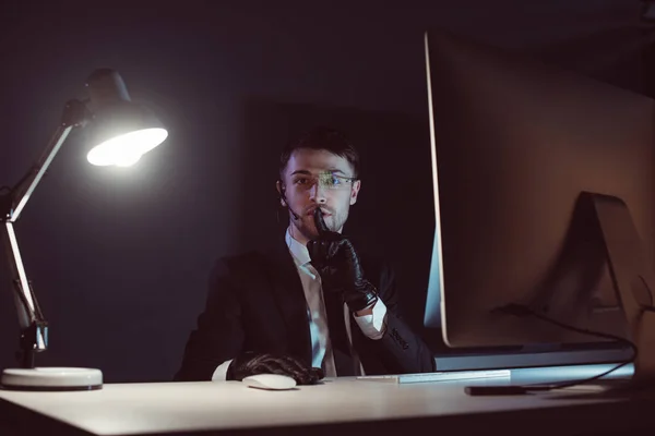 Portrait of spy agent showing silence sign at table with computer screen in dark — Stock Photo