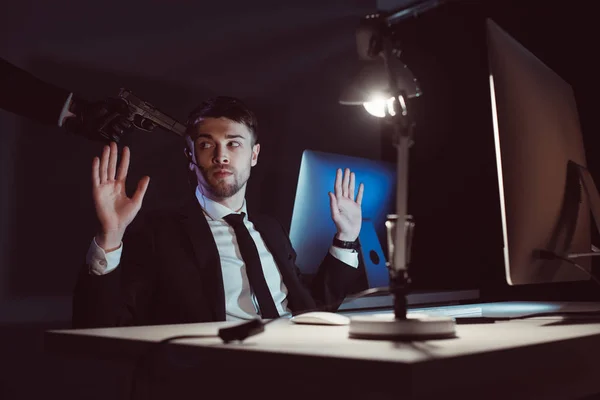 Agent with gun at head and hands up sitting at table with computer screen in dark — Stock Photo