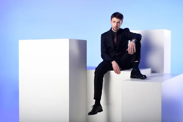 Geschäftsmann im schwarzen Anzug sitzt auf weißem Block isoliert auf blauem — Stockfoto