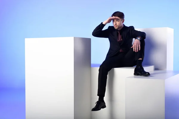 Hombre de negocios en traje negro sentado en el bloque blanco y mirando hacia otro lado aislado en azul - foto de stock