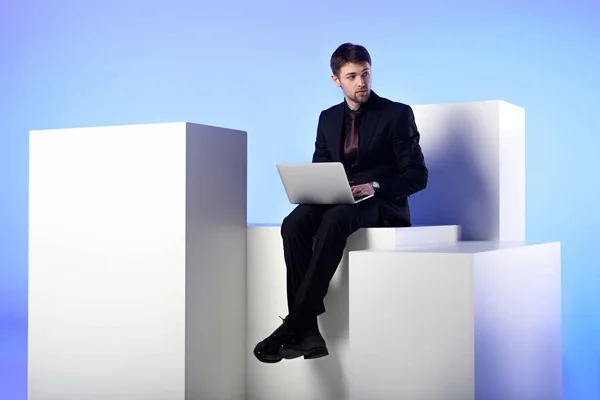 Hombre de negocios con portátil sentado en bloque blanco aislado en blanco - foto de stock