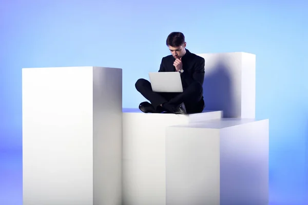 Hombre de negocios usando el ordenador portátil mientras está sentado en el bloque blanco aislado en blanco - foto de stock