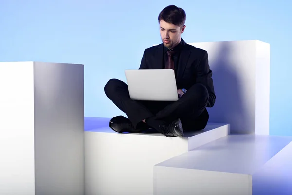 Businessman using laptop while sitting on white block isolated on white — Stock Photo