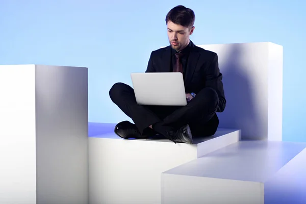 Hombre de negocios usando el ordenador portátil mientras está sentado en el bloque blanco aislado en blanco - foto de stock