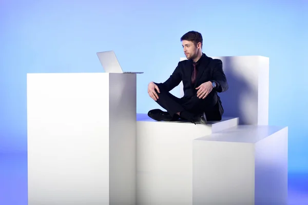 Geschäftsmann mit Laptop sitzt auf weißem Block isoliert auf weißem Block — Stockfoto