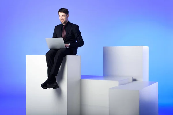 Smiling businessman with laptop sitting on white block isolated on white — Stock Photo