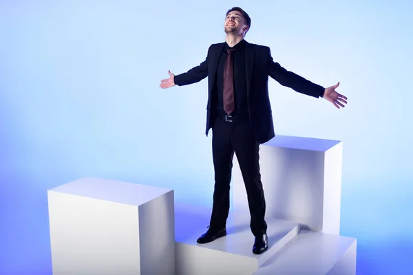 Businessman in black suit with outstretched arms standing on white block isolated on blue — Stock Photo