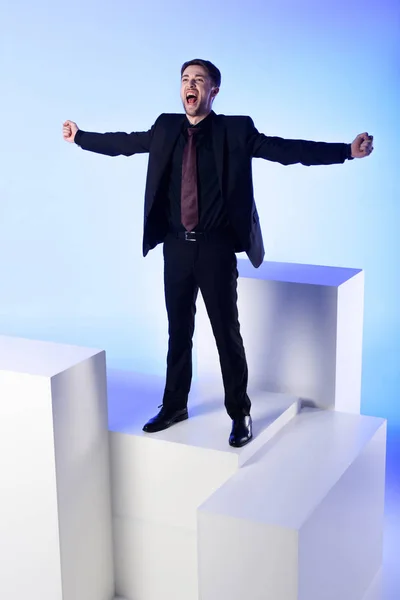 Homem de negócios em terno preto com braços estendidos em pé no bloco branco isolado em azul — Fotografia de Stock