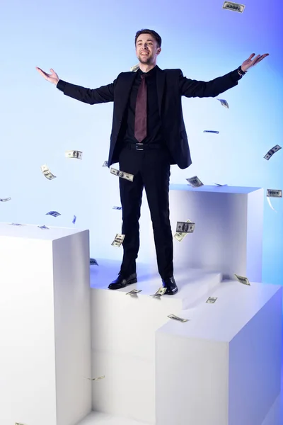 Uomo d'affari sorridente in giacca e cravatta in piedi su blocco bianco con banconote in dollari in caduta intorno isolato su bianco — Foto stock