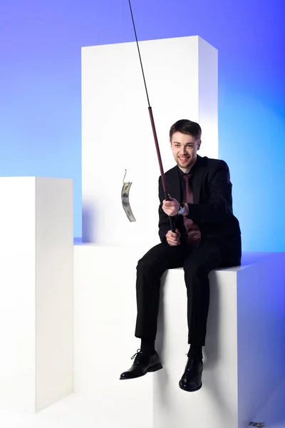 Smiling businessman holding fishing tackle with dollar while sitting on white block isolated on blue — Stock Photo