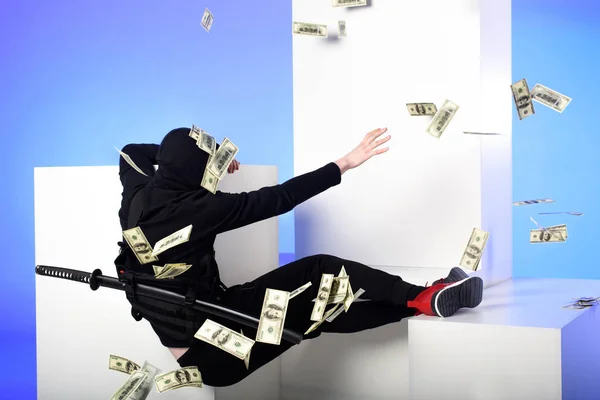 Back view of ninja in black clothing catching dollar banknotes on white blocks isolated on blue — Stock Photo