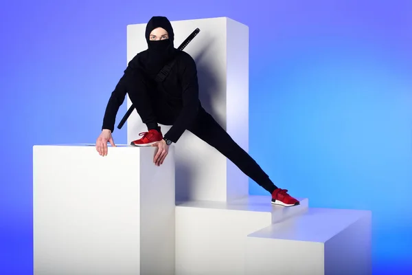 Ninja in schwarzer Kleidung mit Katana dahinter auf weißem Block isoliert auf blauem — Stockfoto