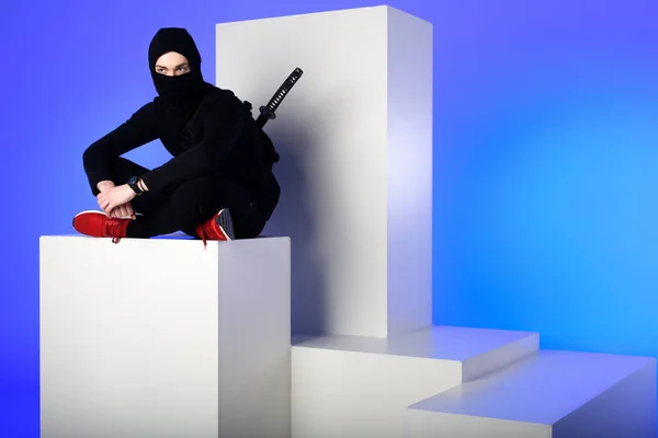 Ninja in black clothing with katana behind sitting on white block isolated on blue — Stock Photo