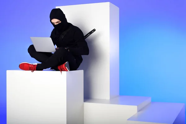 Ninja in black clothing using laptop while sitting on white block isolated on blue — Stock Photo