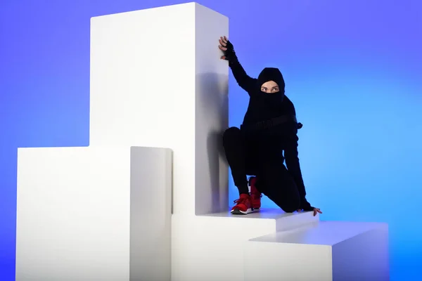 Ninja en ropa negra con katana detrás de pie en bloque blanco aislado en azul - foto de stock