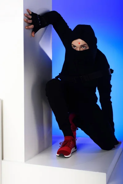 Ninja en vêtements noirs avec katana derrière debout sur bloc blanc isolé sur bleu — Photo de stock