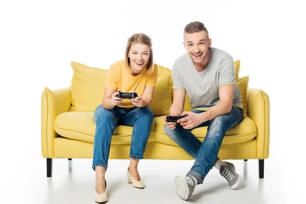 Cheerful couple with joysticks playing video game while sitting together on yellow sofa, isolated on white — Stock Photo