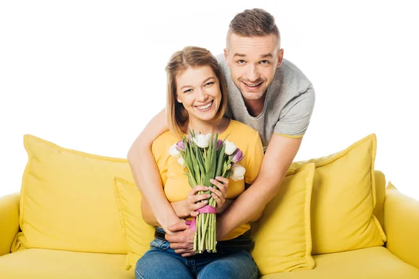 Портрет чоловіка обіймає усміхнену дружину з букетом тюльпанів на жовтому дивані ізольовано на білому — стокове фото