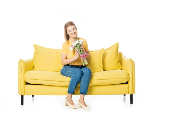 Красива усміхнена жінка з букетом тюльпанів, що сидить на жовтому дивані ізольована на білому — стокове фото