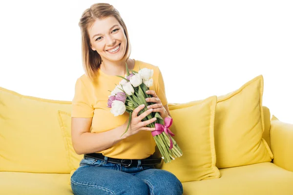 Retrato de hermosa mujer sonriente con ramo de tulipanes sentados en sofá amarillo aislado en blanco - foto de stock