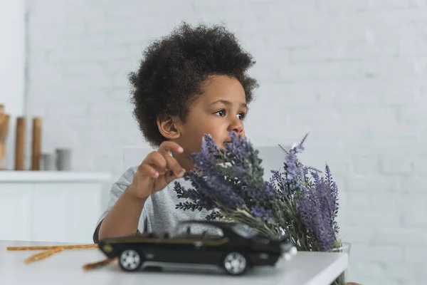 Adorable afroamericano chico tocando violeta flores y mirando lejos en casa - foto de stock